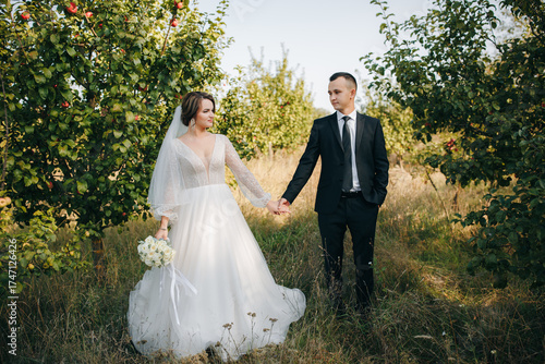Wedding couple holding hands and looking at each other outdoors. Bride in elegant white dress with bouquet and groom in black suit standing among green trees on a sunny day. Love and romance concept.
