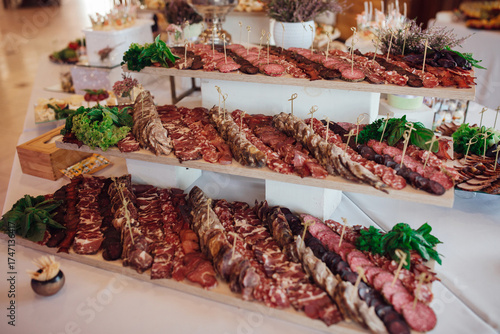 Large assortment of meat appetizers served on multi-level wooden stands. Various sausages, ham and cured meats decorated with greens create an appetizing festive display.