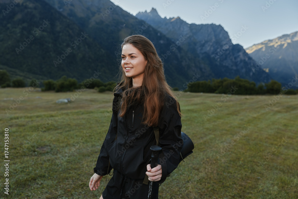 Naklejka premium A young woman in outdoor gear stands in a sunlit grassy field with rugged mountains in the distance, smiling softly as she holds a trekking pole and prepares for adventure.