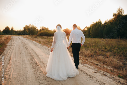 Bride and groom walking along a country road at sunset. The bride wears a beautiful white dress, the groom in a classic suit. Romantic wedding photo shoot outdoors with warm light and genuine emotions
