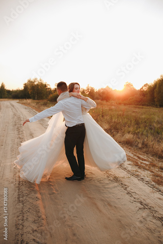 Groom twirls the bride on a sandy road at sunset. Wedding photo shoot in nature capturing motion, joy, and tenderness. Romantic atmosphere with warm evening sunlight and true love.