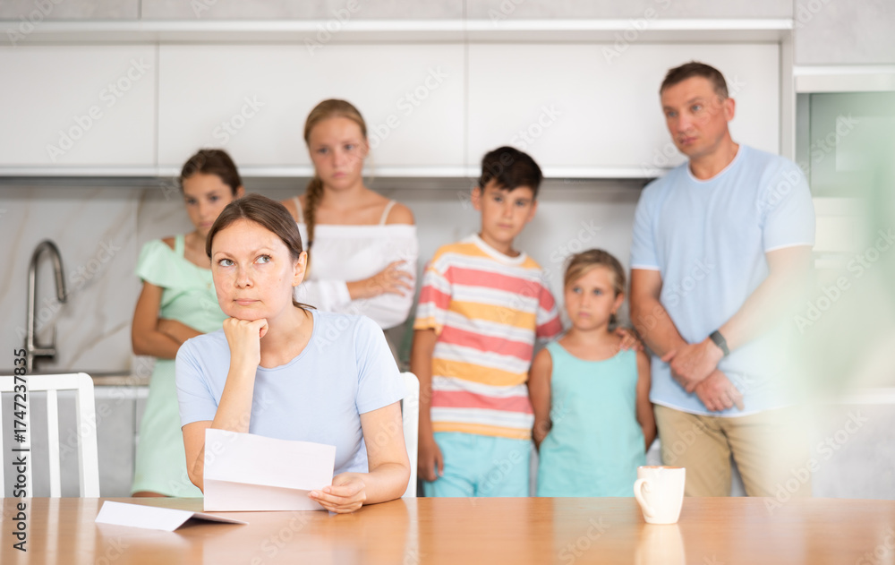 Fototapeta premium Upset mother reads and looks through financial documents. Sad wife with children in the background