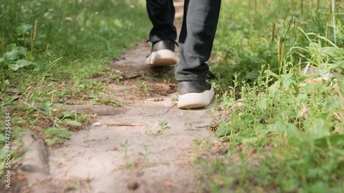 Back leg view of man in black trousers and canvas sneakers walking calmly on forest pathway surrounded by green plants, grass, and soil in peaceful natural outdoor environment during daylight