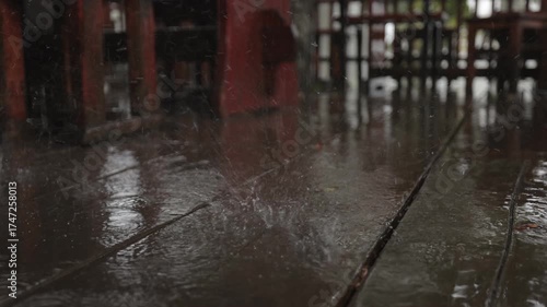 A soothing slow-motion video showing raindrops splashing on a wet wooden deck, forming ripples and reflections. The close-up view captures the calm rhythm and texture of rainfall in detail.