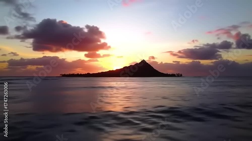 A lone island with a conical mountain is silhouetted against a vibrant sunset sky with colorful clouds.