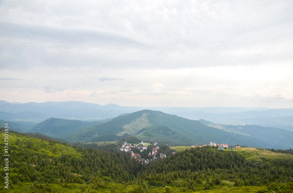 Fototapeta premium Panoramic view of rolling green mountains under a cloudy sky. highest ski resort Dragobrat of colorful roofs is nestled in a valley, surrounded by dense trees. Ukrainian Carpathians