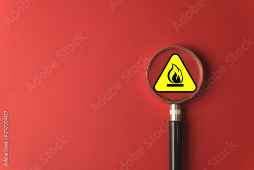Fire risk assessment and hazard identification concept. A magnifying glass focuses on a flammable material warning sign on a red background, symbolizing inspection for potential fire dangers.