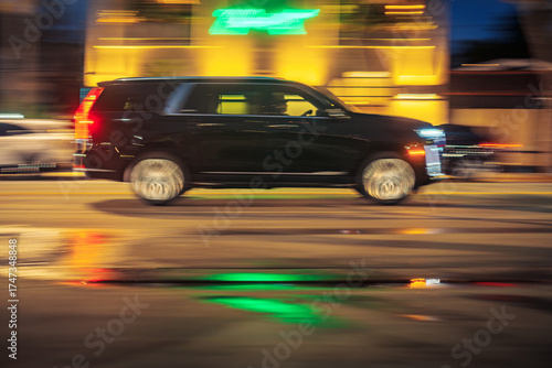 Black executive limo SUV car driving along illuminated wet street at night. Long exposure. Bright colorful neon city lights blurred in background