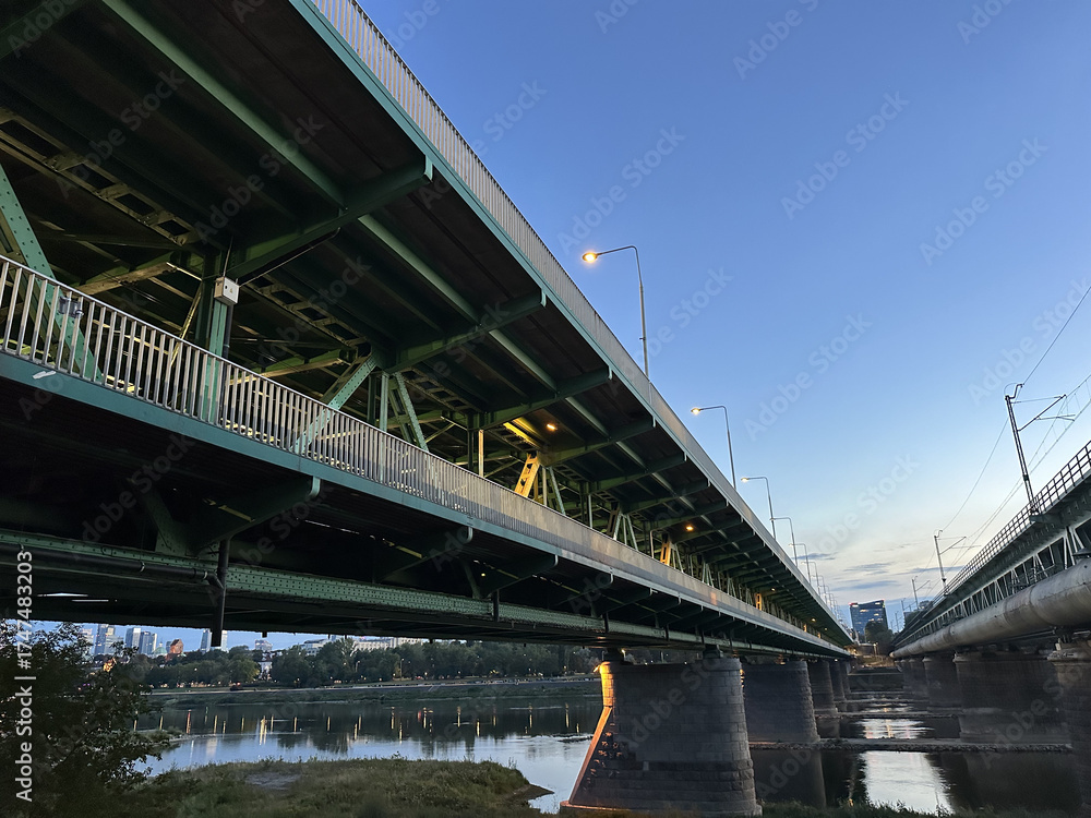 Obraz premium Warsaw, Poland - September 24, 2025: Underneath a large green bridge at dusk, showcasing its structural design and reflections on the water below, highlighting urban architecture and engineering