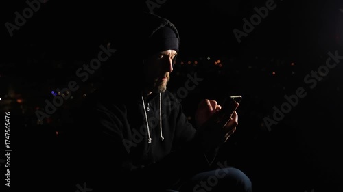 Man in hoodie sitting above Czech city and working on mobile phone at night. Hacker, remote working concept background