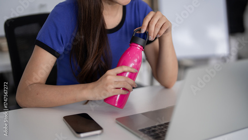 Asian adult woman relaxing at modern office desk staying hydrated with water bottle casual attire enjoying healthy break remote work setup