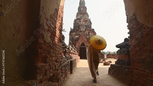 Ayutthaya, Thailand – March 2023: Woman in traditional Thai dress holding yellow umbrella walking by headless Buddha statues at Wat Chaiwatthanaram, UNESCO World Heritage Site symbol of ancient Siam.