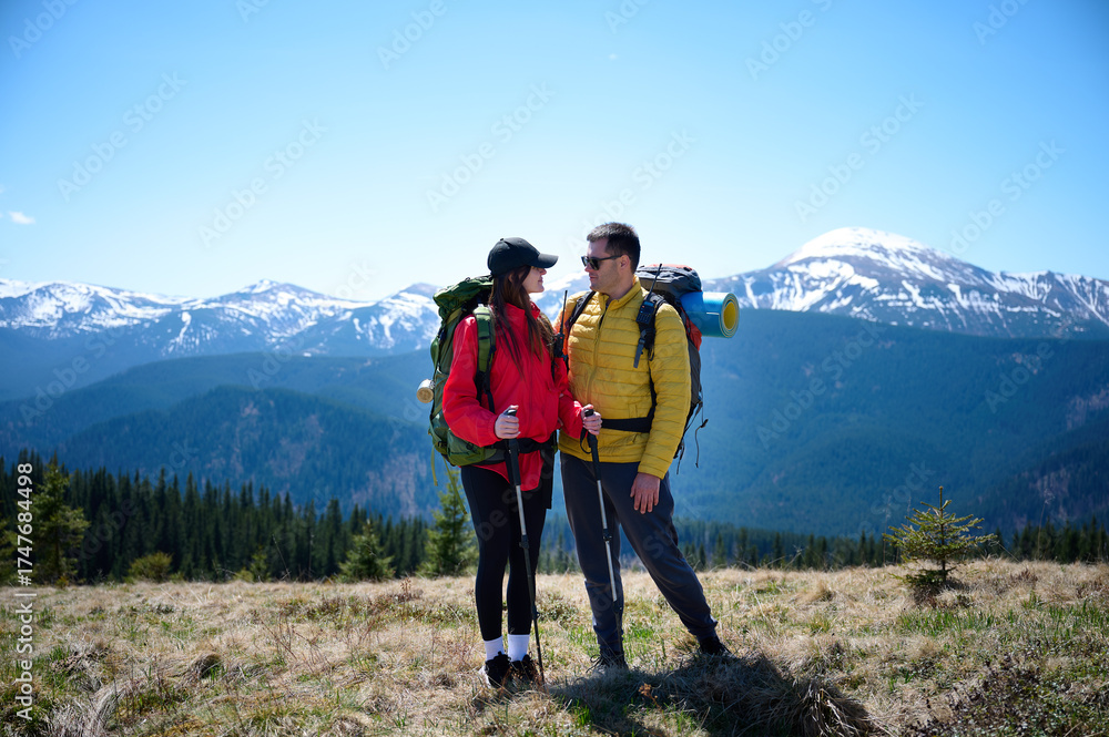 Fototapeta premium Two hikers taking a break on a mountain ridge. Sporty couple with hiking backpacks standing on background of snowy mountains.