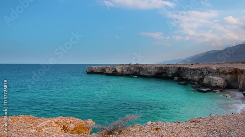 A view of the serene coastline near Qalhat, Oman, showcasing the stunning contrast between the clear turquoise waters of the Arabian Sea and the rugged, rocky cliffs.