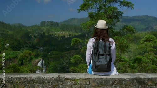 Female traveler sitting on stone wall, gazing at cascading Ramboda Falls surrounded by lush green hills, capturing scenic landscape beauty in tropical Sri Lanka