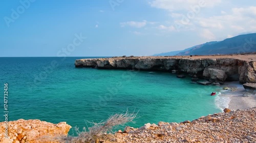 aerial view of the beautiful Fins Beach in Oman, featuring a secluded cove with crystal clear turquoise water and a white sand beach. The rugged, rocky cliffs and dramatic coastline are set against a 