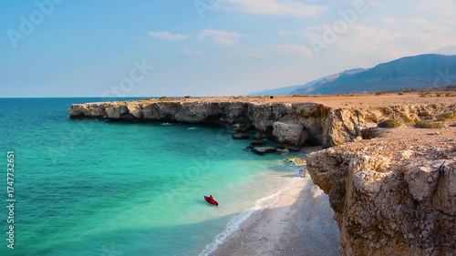 A view of a secluded beach along the Omani coastline, featuring turquoise water, rocky cliffs, and a small red kayak. The serene landscape captures the natural beauty and rugged terrain of Oman's shor
