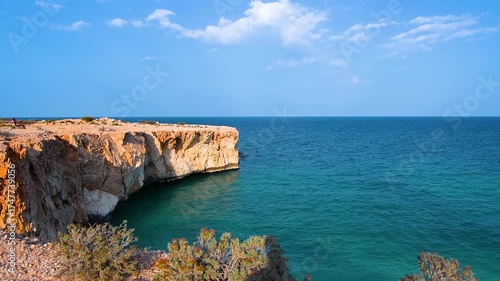 panoramic view of the rugged coastline at Tiwi, Oman. The scene features dramatic, sun drenched rocky cliffs meeting the crystal-clear turquoise waters of the Arabian Sea under a bright blue sky with 