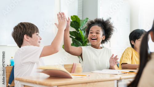 Two cheerful students caucasian and black sit at desks in classroom, smiling giving high five while studying together with notebooks open. Education back to school