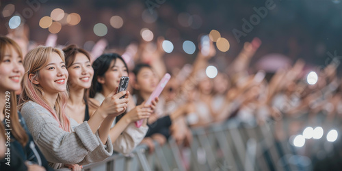 A group of joyful young Korean women with blonde and pink hair in the front row of an idol concert, smiling and recording the show on their phones amidst a cheering audience of fans.