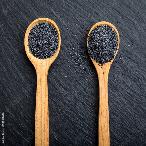 Two wooden spoons filled with dark poppy seeds, on a textured black slate