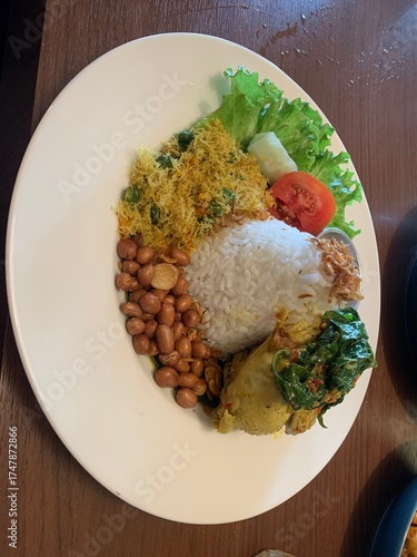 Balinese dish served on white plate featuring Bebek Betutu roast duck, accompanied by amound of rice topped with fried shallots. includes sauteed greens contain grated coconut, roasted peanuts, salad.
