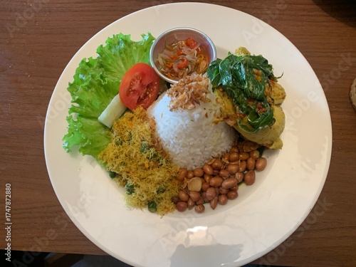 Balinese dish served on white plate featuring Bebek Betutu roast duck, accompanied by amound of rice topped with fried shallots. includes sauteed greens contain grated coconut, roasted peanuts, salad.