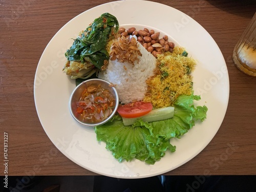 Balinese dish served on white plate featuring Bebek Betutu roast duck, accompanied by amound of rice topped with fried shallots. includes sauteed greens contain grated coconut, roasted peanuts, salad.