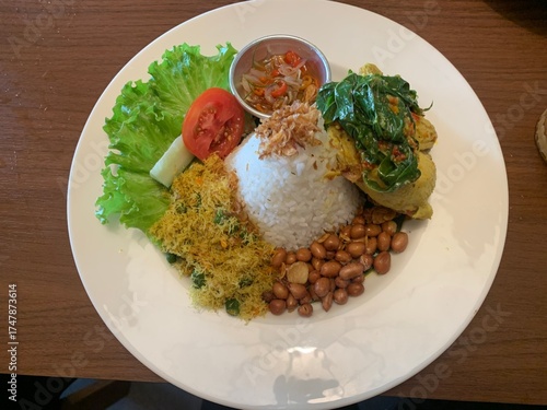 Balinese dish served on white plate featuring Bebek Betutu roast duck, accompanied by amound of rice topped with fried shallots. includes sauteed greens contain grated coconut, roasted peanuts, salad.