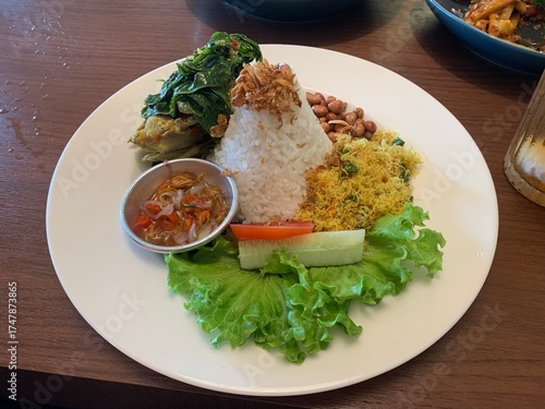 Balinese dish served on white plate featuring Bebek Betutu roast duck, accompanied by amound of rice topped with fried shallots. includes sauteed greens contain grated coconut, roasted peanuts, salad.