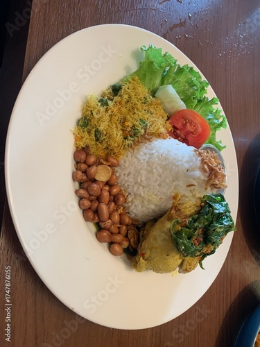 Balinese dish served on white plate featuring Bebek Betutu roast duck, accompanied by amound of rice topped with fried shallots. includes sauteed greens contain grated coconut, roasted peanuts, salad.