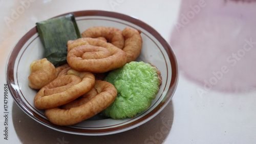 Assorted Indonesian traditional snacks on a plate, featuring fried spiral pastries, pandan rice cake, and banana leaf-wrapped treat. Ideal for culinary visuals and cultural food themes.