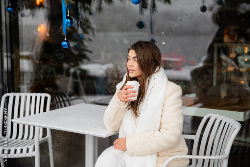 Young woman in a white winter coat and scarf sitting at an outdoor café with a takeaway coffee. Snow and blue Christmas ornaments create a cozy festive atmosphere.