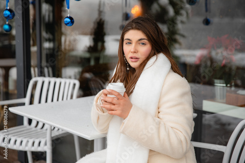 Young woman in a white winter coat and scarf sitting at an outdoor café with a takeaway coffee. Snow and blue Christmas ornaments create a cozy festive atmosphere.