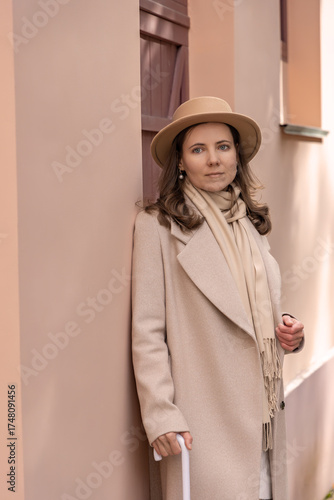 Refined portrait of a woman in a beige coat, scarf and hat leaning by a pastel wall with soft natural light