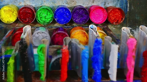 On a wooden workbench in a screen printing shop, several plastisol ink cans are open, revealing rich tones soft pastels, strong primaries, and deep shades. A wooden spoon and trowel rest nearby.