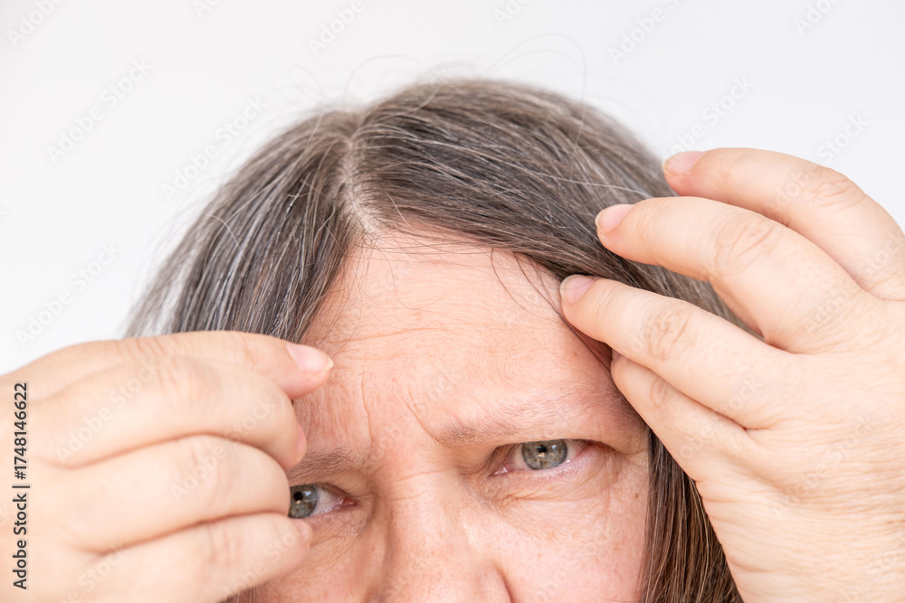 Fototapeta premium Mature woman touching her gray hair, examines silver strands, deep in thought, aging process, menopause time, emotional reflection, quiet acceptance