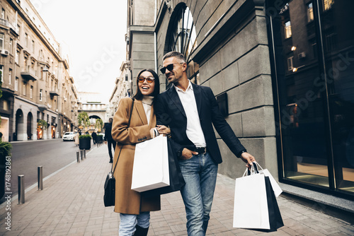 Fashionable couple walking in city with shopping bags, laughing and enjoying time together. Man in black jacket and white shirt,  Asian woman in camel coat and sunglasses. Black friday sales.