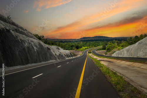 Quadro em tela A long, straight road stretches into the distance, leading towards a mountain range under a warm, orange sky