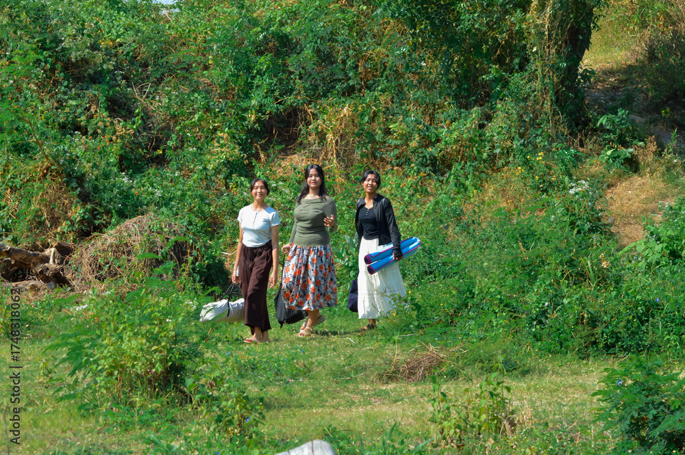 Fototapeta premium Three girls stroll under bright sunlight across a grassy clearing, each carrying bags and mats as they look for a good spot to rest and start their outdoor camp.