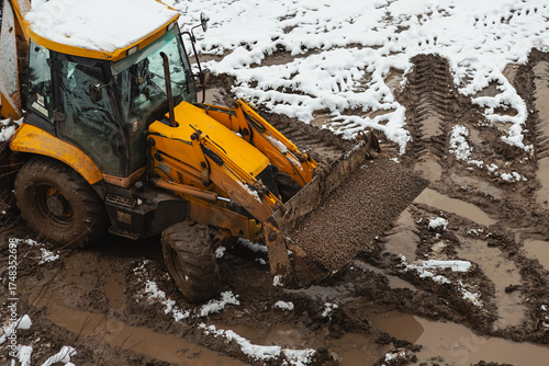 The bucket of a loader tractor is moving along a dirty dirt construction road in winter. Construction during the winter season.