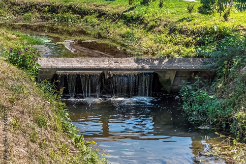 Concrete waterfall weir with flowing water and lush vegetation at Kozatska Prystan recreation base in Romny, Sumy region, Ukraine. Peaceful natural stream landscape with reflections
