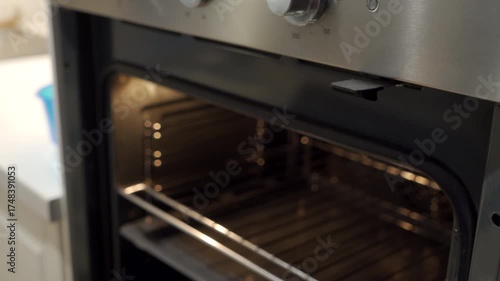 A hand, with fingers in motion and a reddish hue, grasps the metallic handle of an oven door. The oven has a brushed stainless steel finish and the door is slightly ajar, revealing a dark interior.