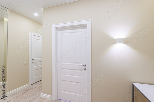 A hallway with neutral walls features a white panel door, illuminated by a modern sconce. Another door leads into the space, with a glimpse of flooring and a mirror on the left