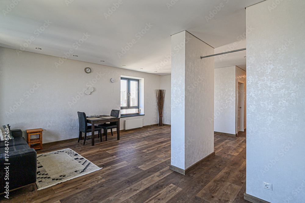 Fototapeta premium Bright, modern living space with wood floors. A dining table sits by a window, and a black sofa is to the left. White walls and textured pillars add visual interest