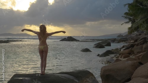 A woman enjoys a peaceful moment in slow motion on a Seychelles beach at sunrise or sunset, gently dancing on a rock by the sea with her hands raised toward the sky.
