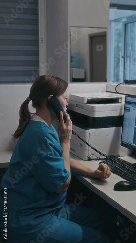 Vertical shot of Caucasian female medical assistant in blue scrubs picking up phone and booking appointment while working on computer in private clinic