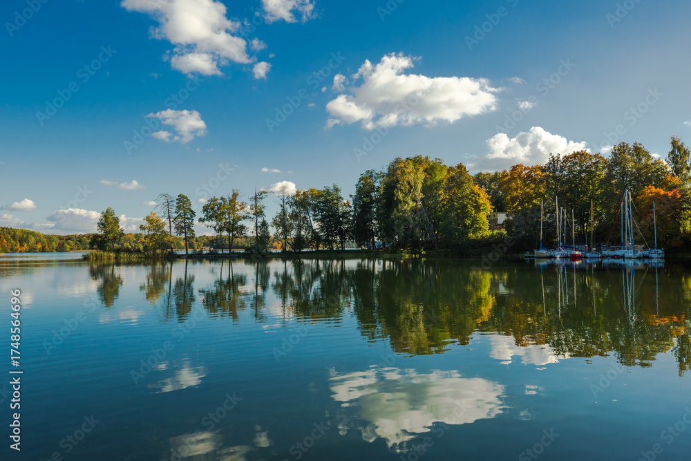 Obraz premium A picturesque autumn landscape on the shore of a lake. A line of trees rises above the smooth water, reflecting a blue sky with white clouds. Several sailing yachts are moored near the shore.