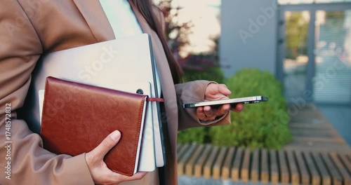 Close-up of woman's hands holding laptop and notebook in one hand while typing message on her smartphone with the other hand, outside