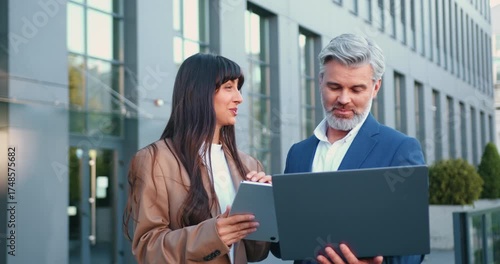 Close-up of mature businessman with laptop and stylish young businesswoman with tablet standing outside modern office building, collaborating on project and discussing business strategy